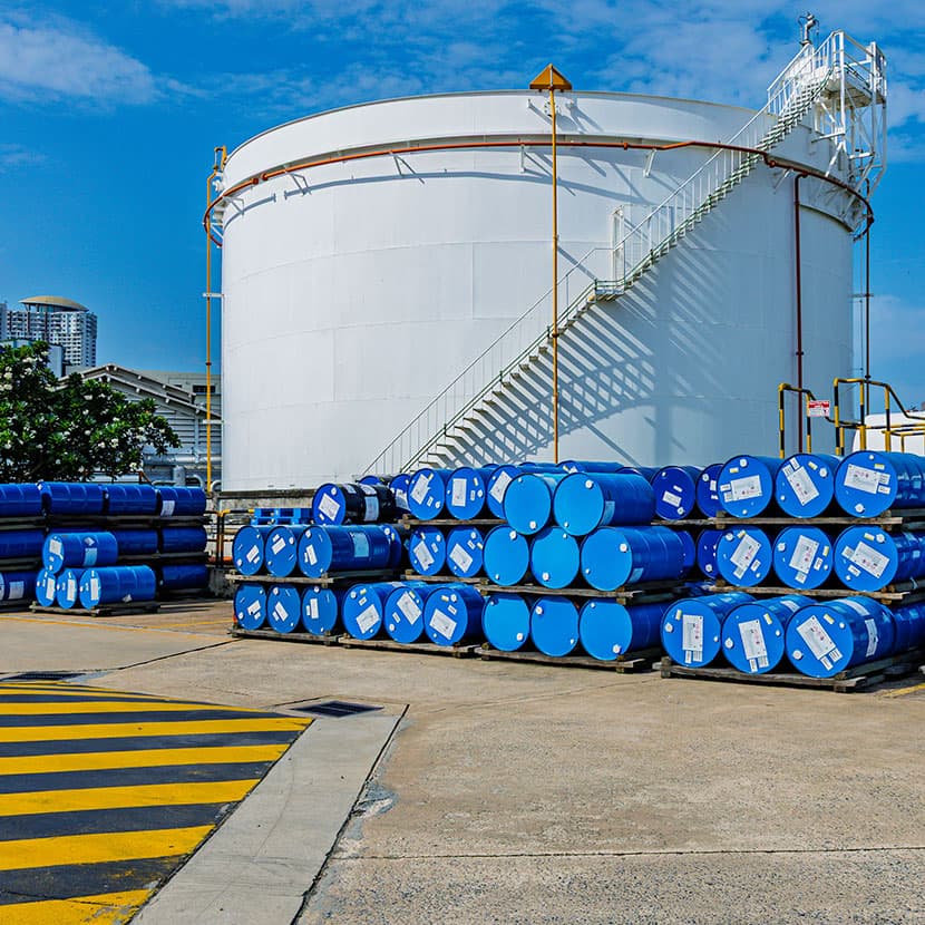 Large industrial storage tank with stacked blue chemical drums in an outdoor facility under clear sky