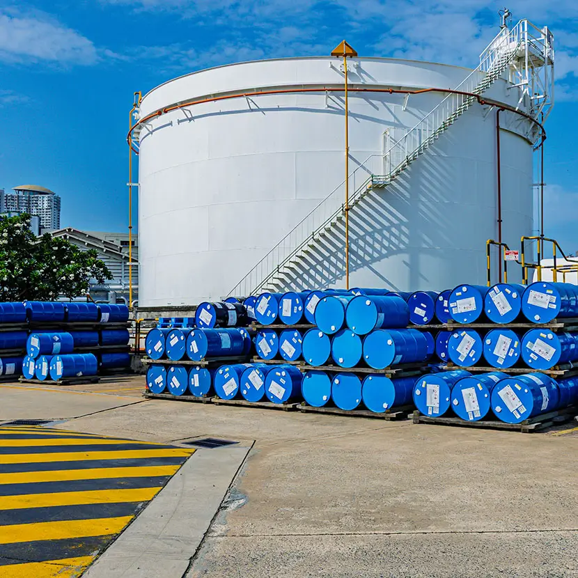 Large industrial storage tank with stacked blue chemical drums in an outdoor facility under clear sky