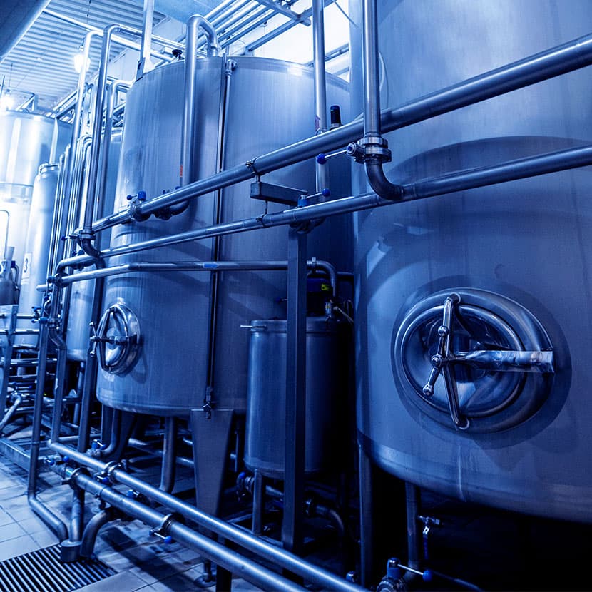 Industrial stainless steel tanks and pipes inside a modern beverage or food processing plant with blue-toned lighting.